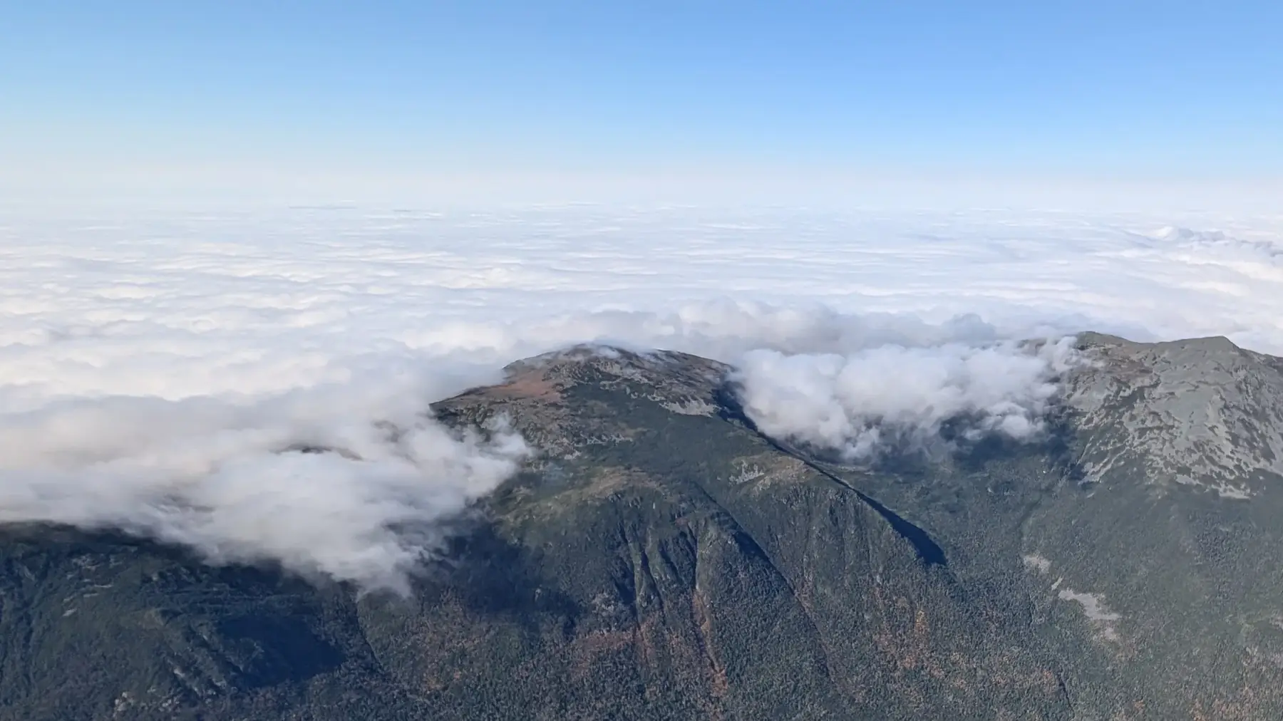 Cloud spilling over a mountain ridge, with an undercast beyond and blue sky above