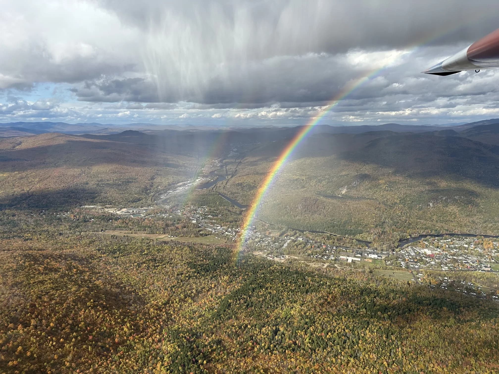 Snow virga falling as a rainbow arches over the river valley below, seen from the wing of a glider